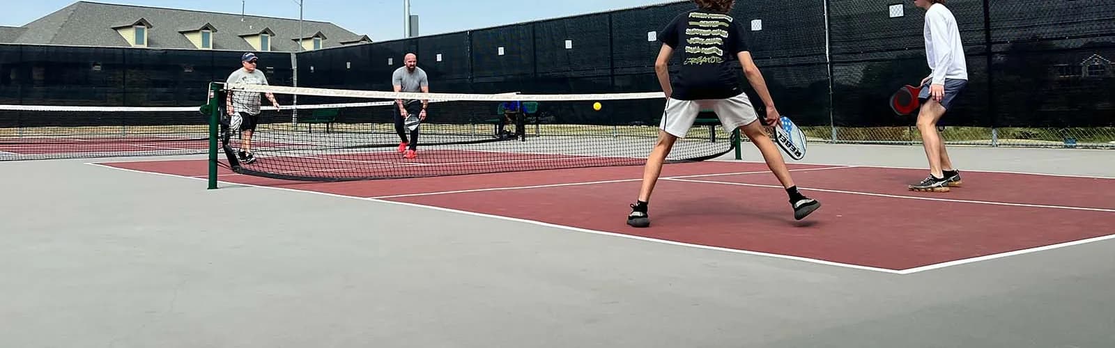 Sunlit outdoor pickleball courts with players in an active doubles rally