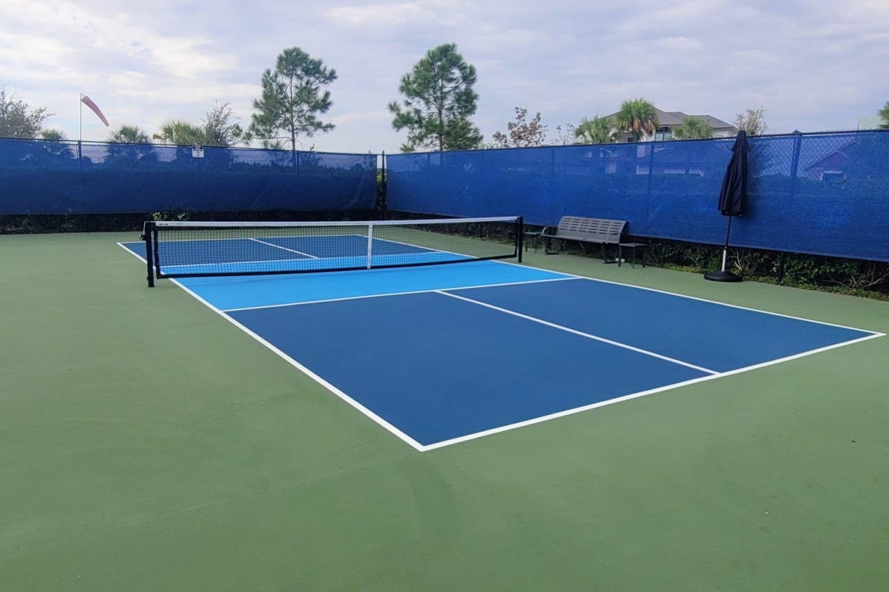 A two-tone blue pickleball court with a bench and pine trees under a partly cloudy sky, stock photo for Daffin Park Tennis Courts in Savannah, GA