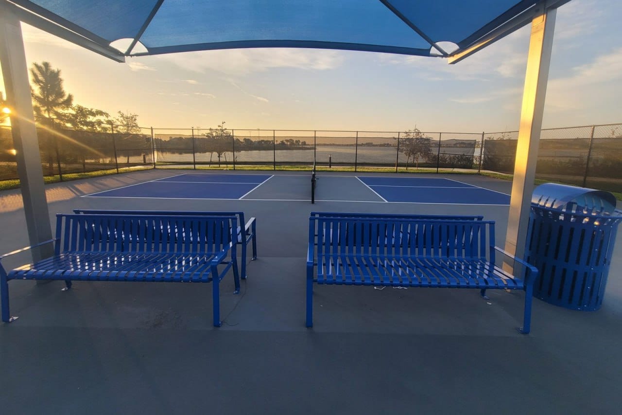 A view from under a blue canopy with benches to a pickleball court beside a lake at sunrise, stock photo for Doral Legacy Park (Doral) in Doral, FL
