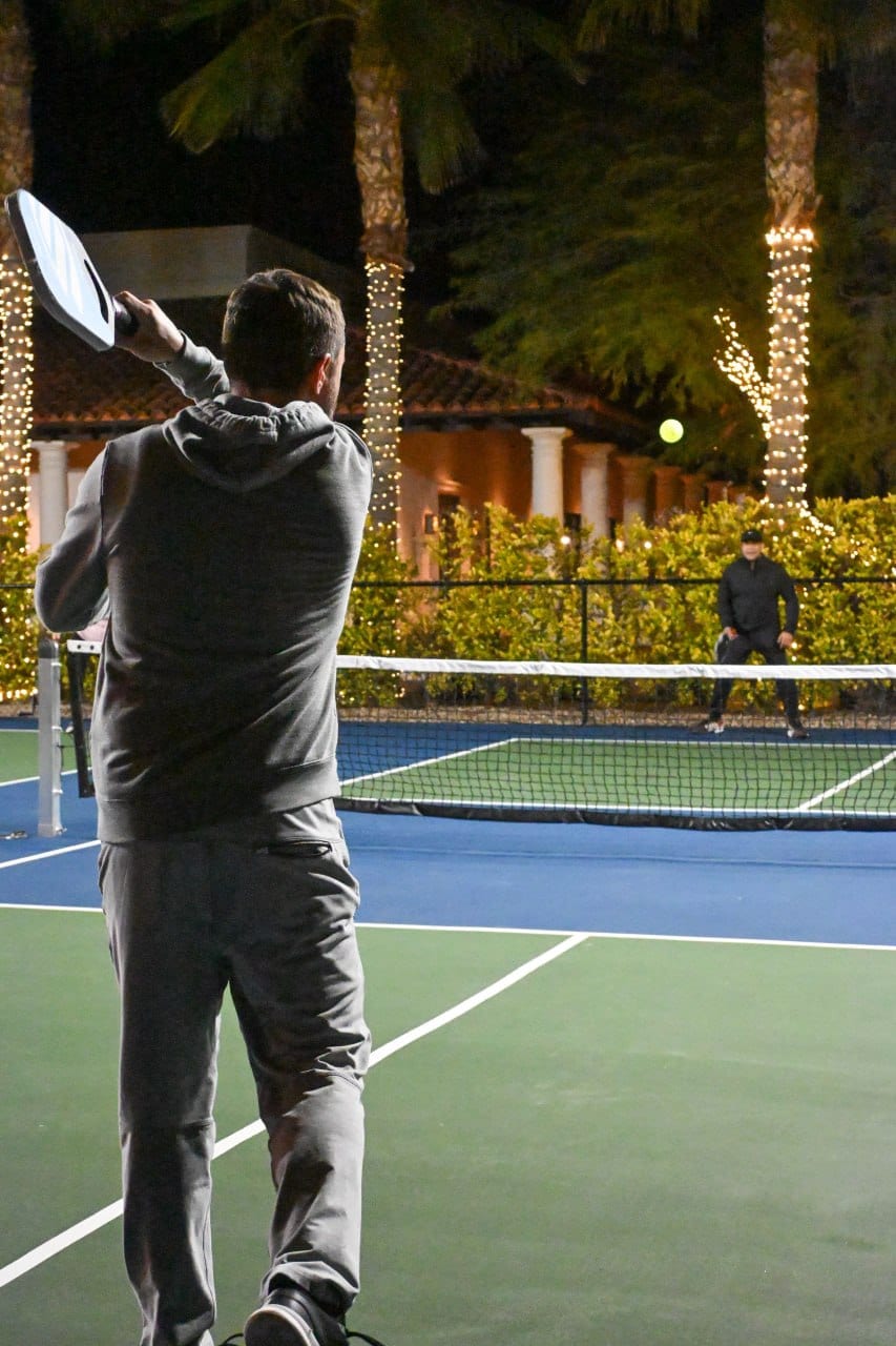 Two players at a night pickleball game under string lights, with palm trees and a columned building behind the court, stock photo for Chatham Racquet Club in Savannah, GA